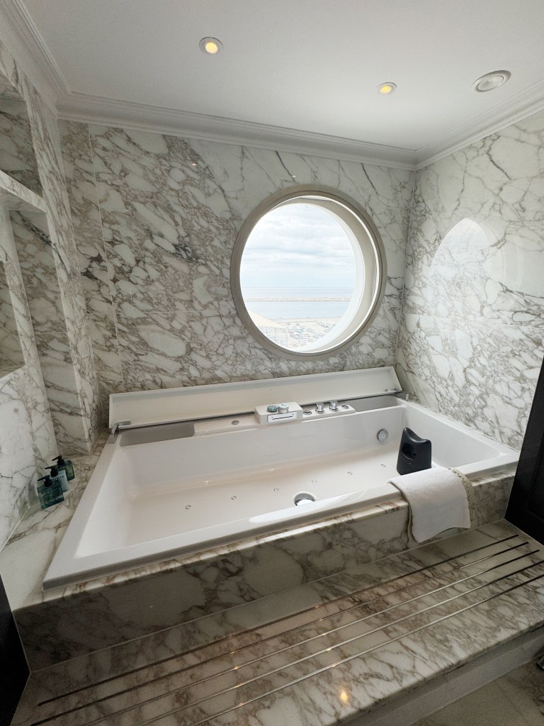 A large marble style bath with a large porthole next to it.