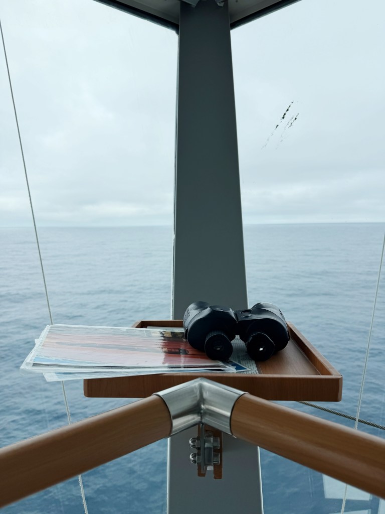 A shelf in the bridge with binoculars on it and a view of the ocean behind.