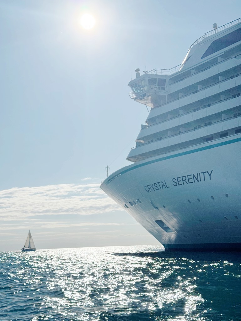 Crystal Serenity Cruise Ship at anchor in the sunshine with a small sailing yacht in front of the ship.