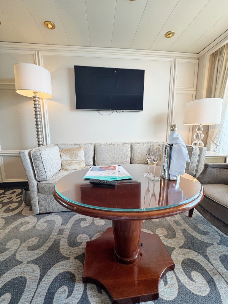A sofa, with a large TV on the wall and a coffee table with a bottle of Champagne and two glasses