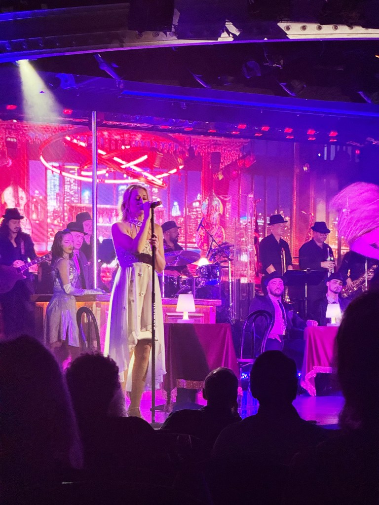 A singer, standing at a microphone with other singers, dancers and musicians in the background.