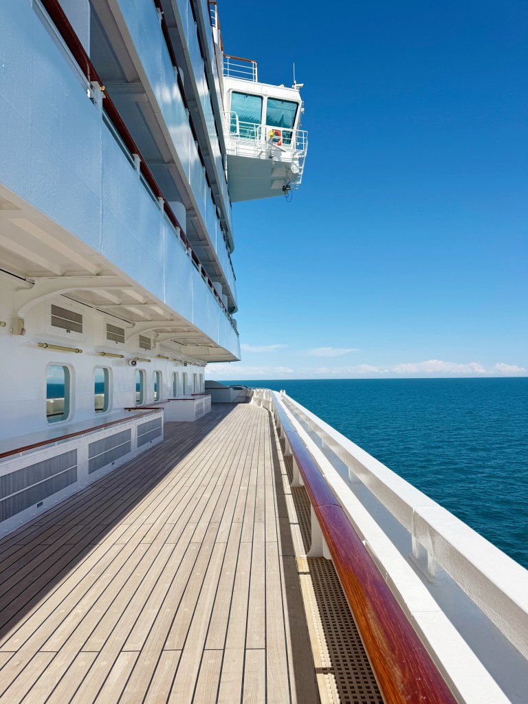 A wide, teak promenade deck whislt Crystal Serenity is at sea, with a view of the Bridge Wing above.