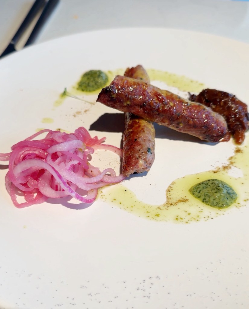 Three grilled Kebabs served on a white plate with green sauce and condiments, accompanied by pickled red onions.