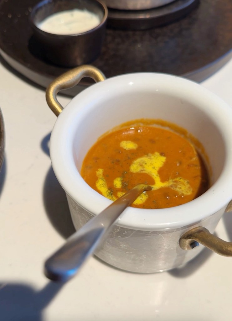 A small white bowl with handles containing a smooth, orange sauce topped with a swirl of cream and herbs, alongside a metal dish of white yogurt sauce.