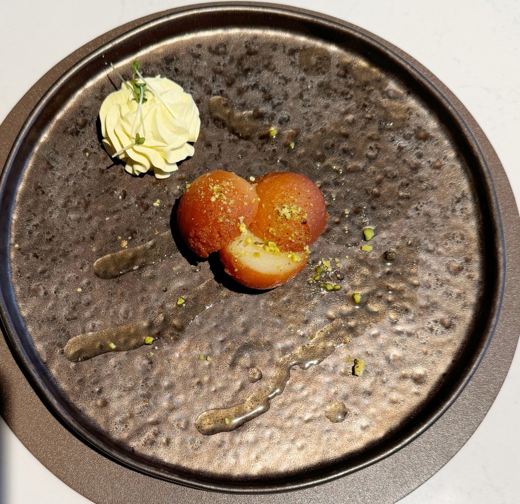 A plate featuring three orange-red spherical desserts, topped with crushed pistachios, accompanied by a dollop of whipped cream on the side.