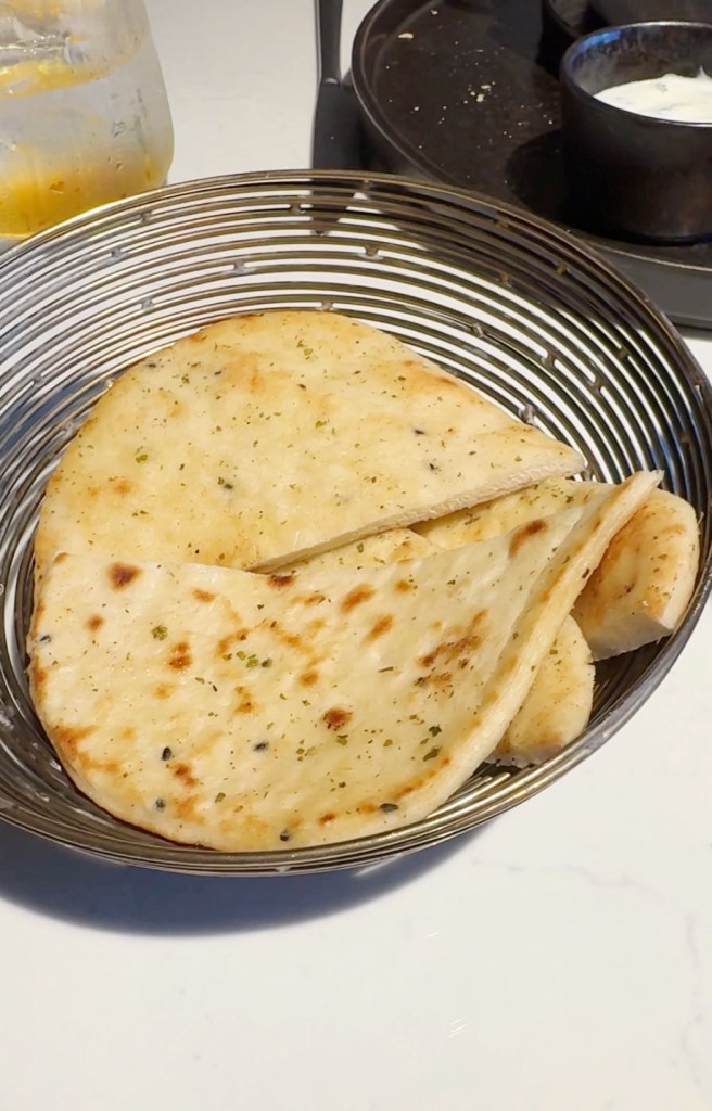 A basket containing several pieces of freshly baked flatbread, some of which are folded, served alongside a glass of drink and small bowls of condiments.