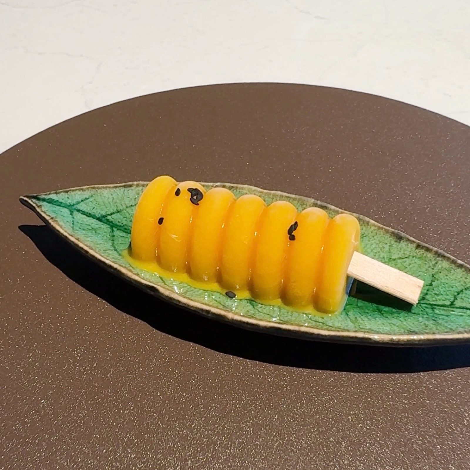 A yellow dessert shaped like a stack of rounded segments, presented on a green leaf-shaped dish with a wooden stick and black flecks.