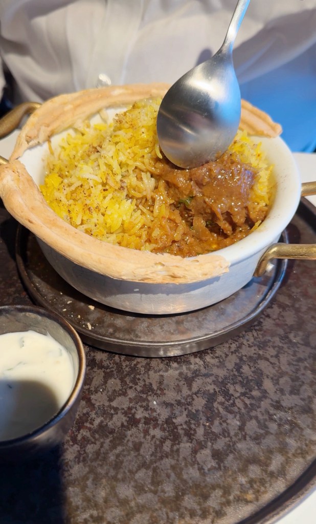 A bowl of yellow rice garnished with spices, alongside a serving of meat curry, with a spoon about to scoop from the dish. A small bowl of yogurt sauce is placed beside it.