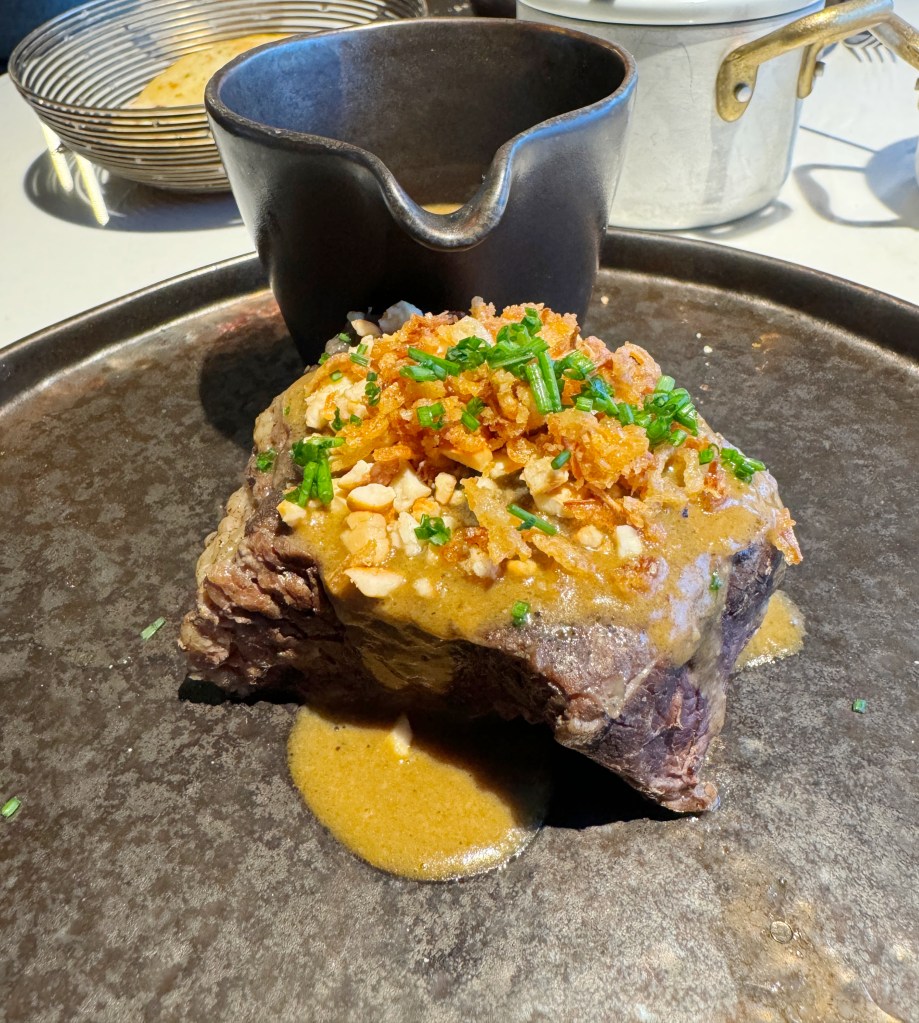 A plated portion of tender beef steak topped with crispy fried onions and chopped chives, accompanied by a small bowl of sauce.