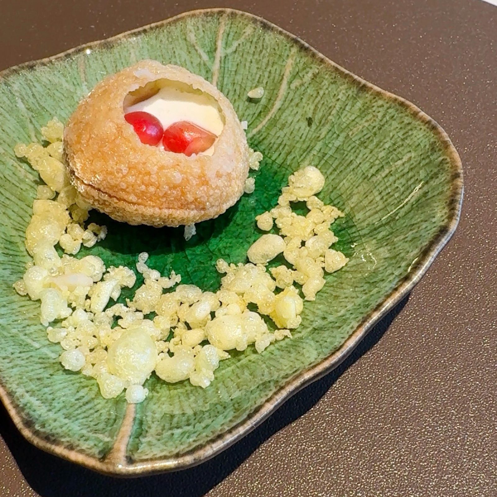 A delicate pre appetiser featuring a hollow, crispy shell filled with yoghurt and garnished with pomegranate seeds, served on a green leaf-shaped plate with crunchy bits around it.