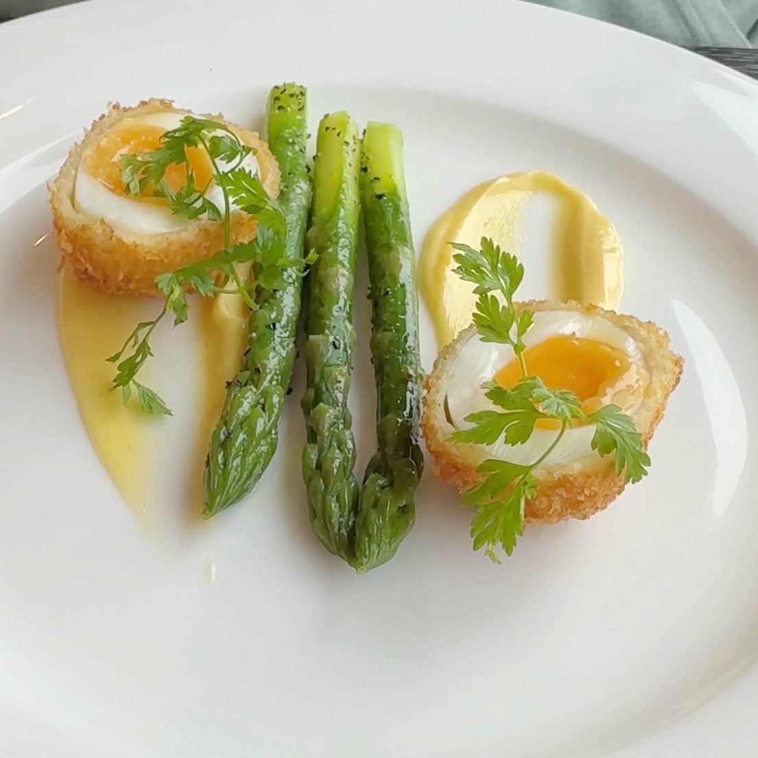 Two halves of a soft poached egg, with some hollandaise sauce, 3 spears of asparagus and some parsley garnish.