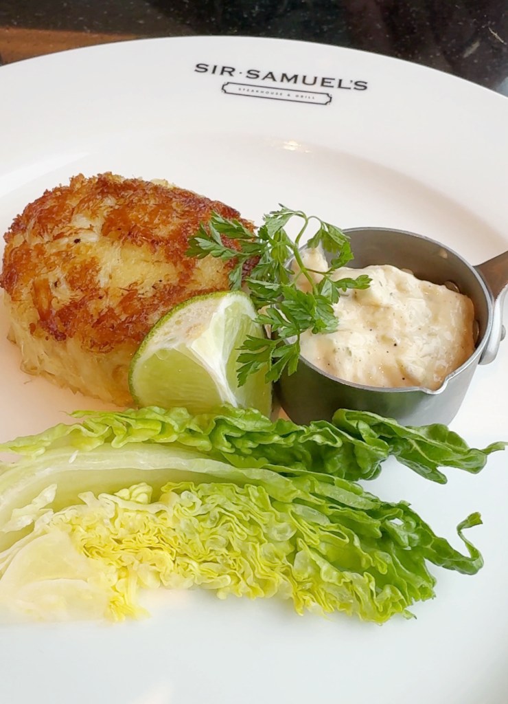 A Jumbo lumb crab cake with lettuce, a wedge of line and some tartar sauce.