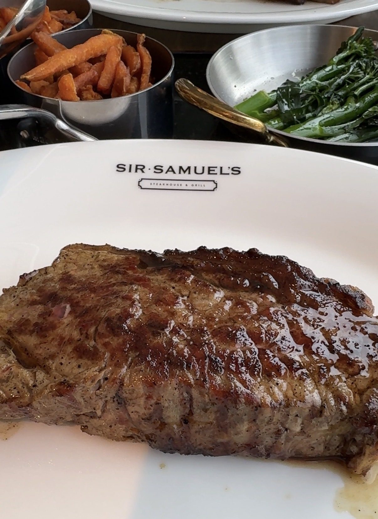 A Steak on a branded Sir Samuel's plate with sides of sweet potato and tenderstem brocolli in dishes behind.