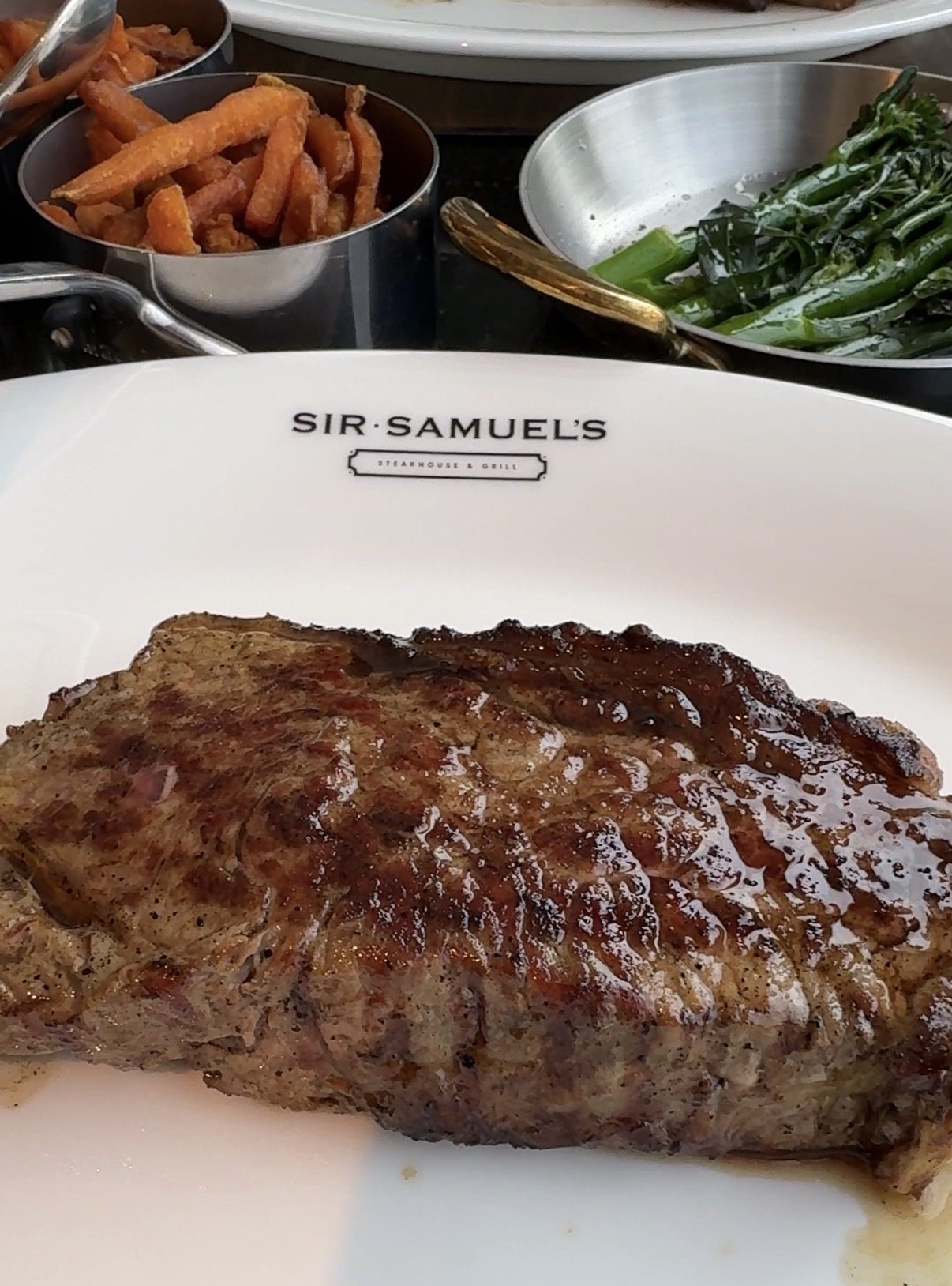 A Sirloin steak on a branded Sir Samuel's plate, with side dishes of sweet potato fries and tenderstem brocolli in the background.